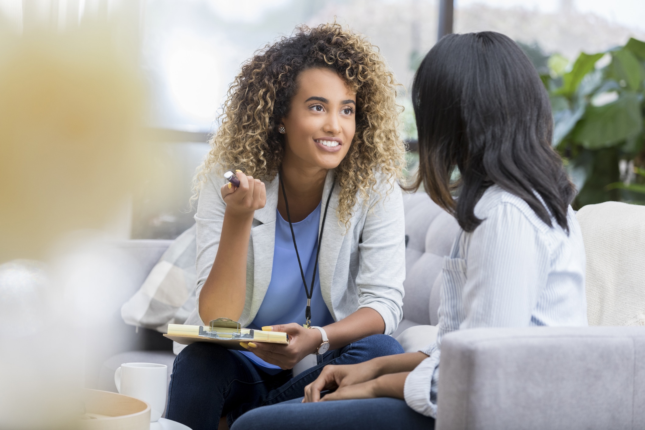 A cheerful young therapist sits on the couch with an unrecognizable client.  She holds a clipboard and gestures as she speaks to her.