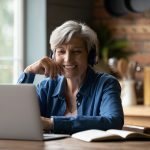 Smiling modern mature 60s Caucasian woman in earphones look at laptop screen watch webinar online at home. Happy senior grey-haired female have fun study on internet. Elderly technology concept.